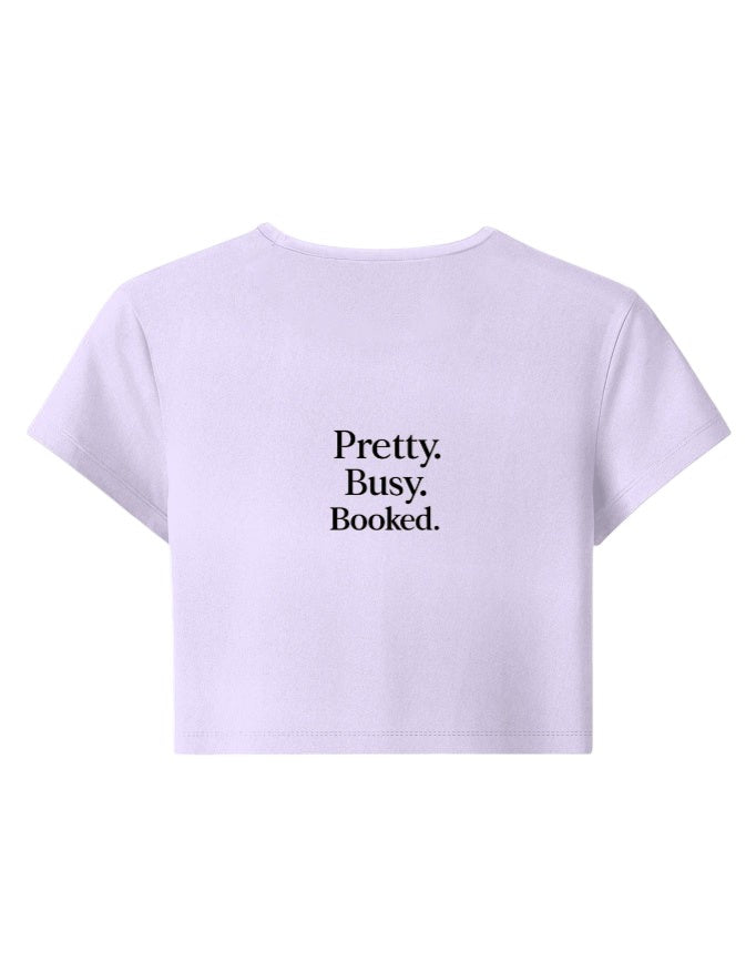 Fourthwave lilac baby tee with black text that reads "Pretty. Busy. Booked.". Cropped, short-sleeve, 95% cotton, 5% spandex.